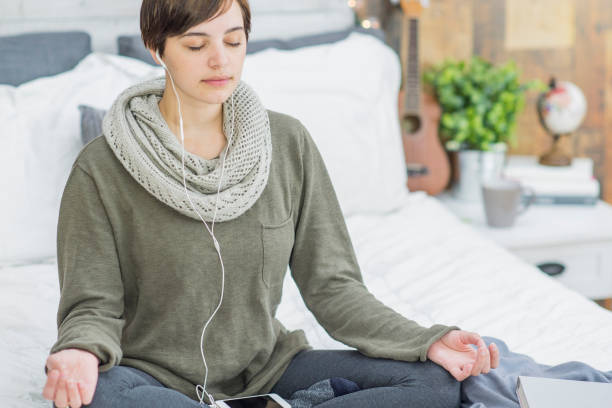 A Caucasian woman is indoors in her bedroom. She is wearing casual clothing. She is sitting on her bed and listening to music on her cell phone. She is wearing headphones and meditating.