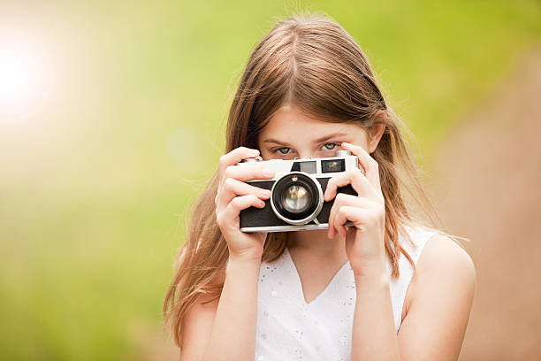 A beautiful 12 year old girl peeking from behind a vintage camera.