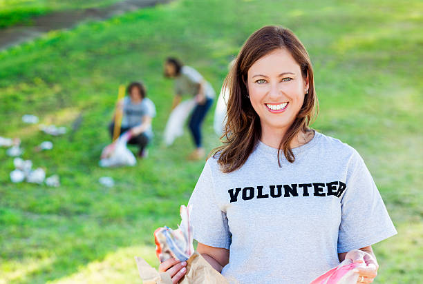 Beautiful Caucasian woman holds trash bag as she picks up trash with volunteer teem in local park. Other volunteers are in the background working.