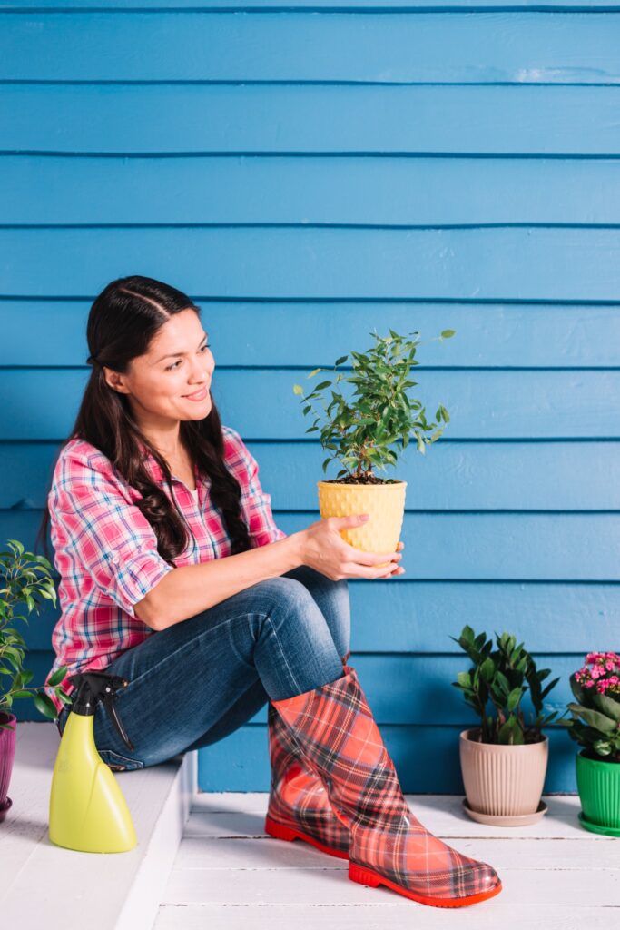 Woman in plaid shirt and red boots holding a potted plant while gardening on a porch with colorful flower pots around her.