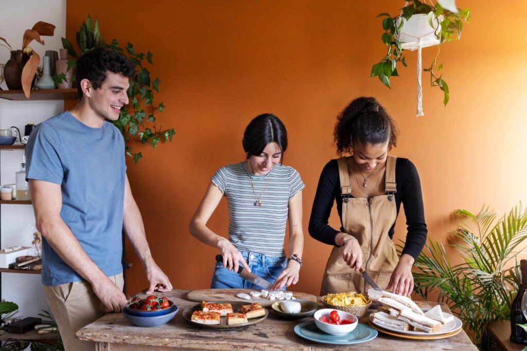 Three friends preparing food together in a cozy kitchen with an orange wall, enjoying a fun cooking session.