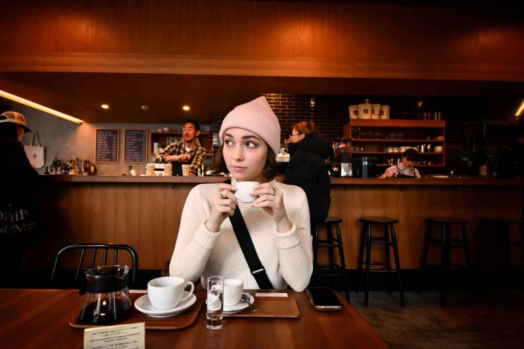 A woman wearing a light pink beanie and cream sweater is sitting at a café table, holding a cup of coffee and looking to the side. Behind her, baristas prepare drinks at a wooden counter with a warm, cozy interior.