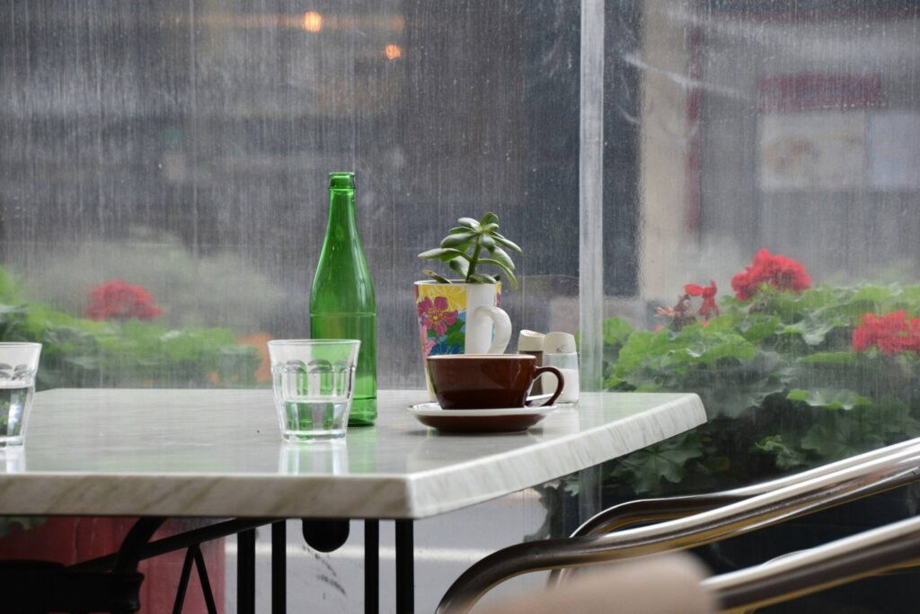 A cozy café table by the window on a rainy day, featuring a coffee cup, glass bottle, and potted plant with raindrops on the glass.