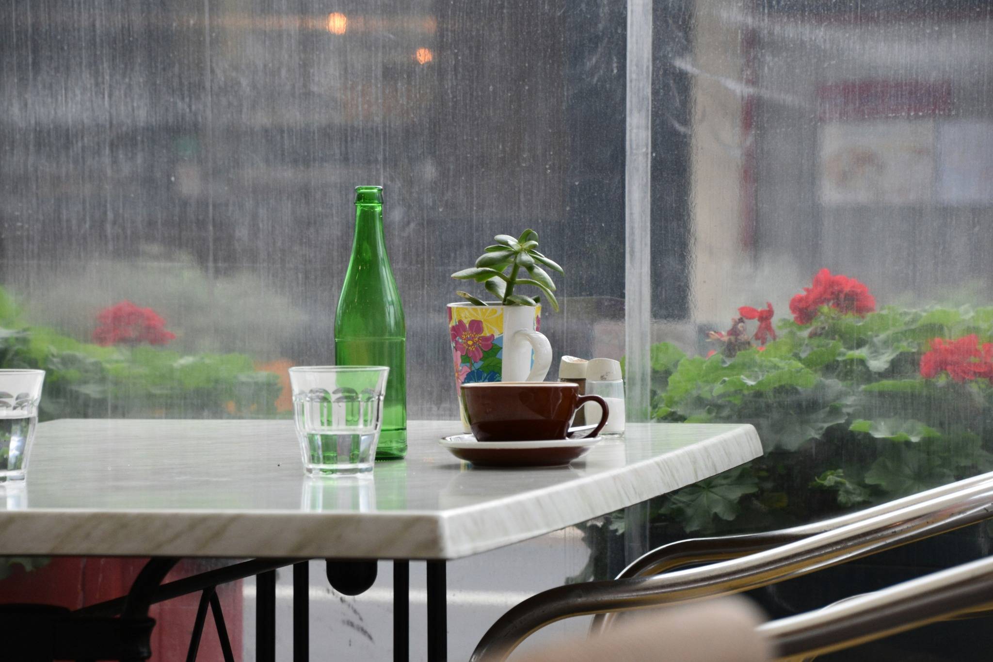 A cozy café table by the window on a rainy day, featuring a coffee cup, glass bottle, and potted plant with raindrops on the glass.