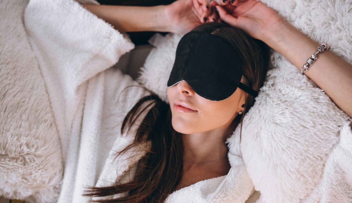 Woman relaxing on a bed while wearing a black eye cover for better sleep and comfort