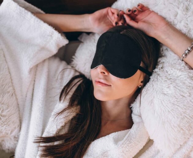 Woman relaxing on a bed while wearing a black eye cover for better sleep and comfort