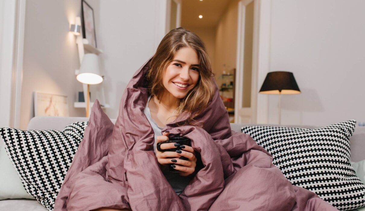 Woman wrapped in a cozy blanket holding a warm drink, enjoying a peaceful evening at home — perfect example of winter self care.